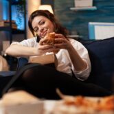 Woman talking at phone with friends while holding tasty delicious burger in hands relaxing on couch in living room late at night. Caucasian female enjoying tajeaway junk food home delivered